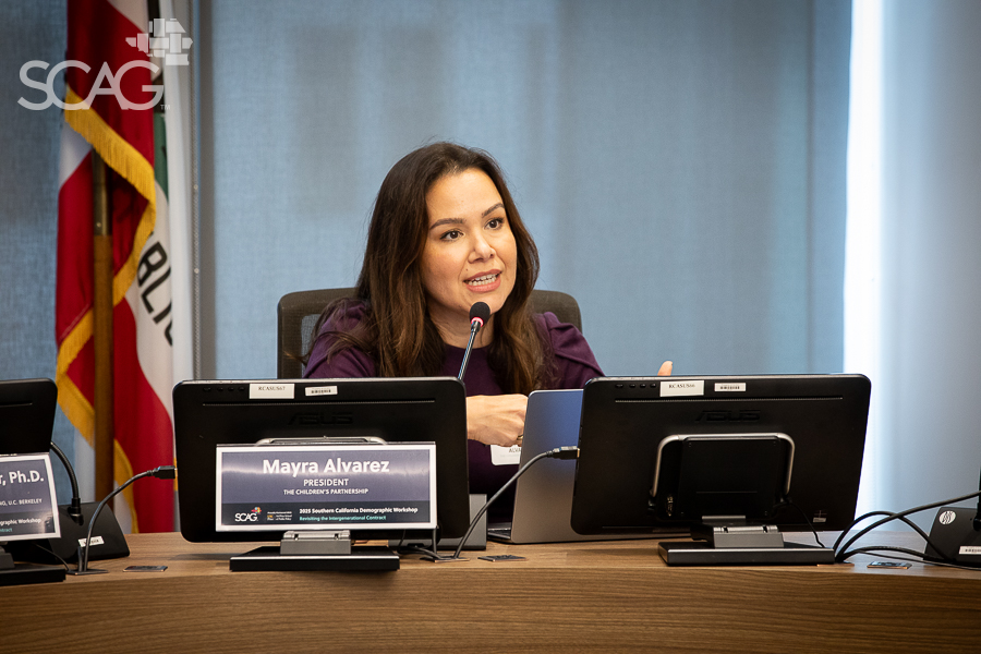 Woman speaking at a conference table with monitors and flags behind.