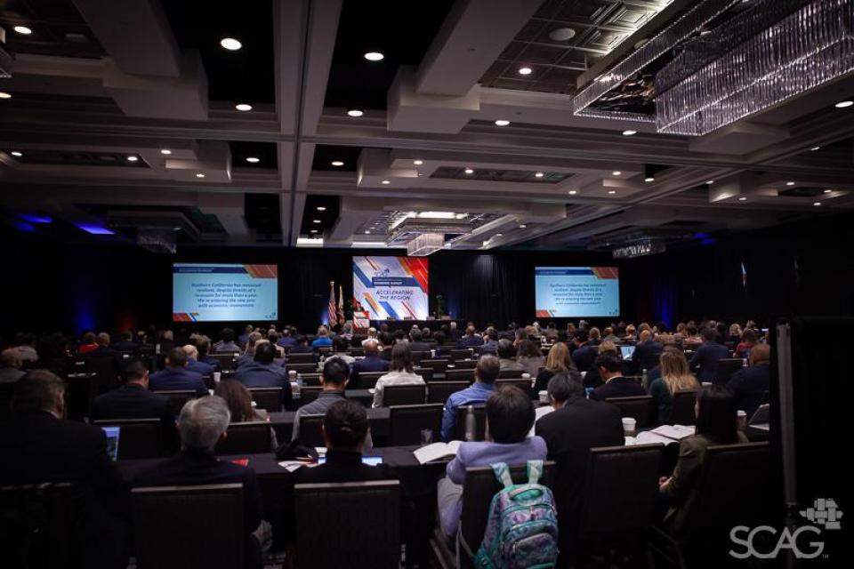Overall room  at the Economic Summit filled with attendees, a stage and two screens.